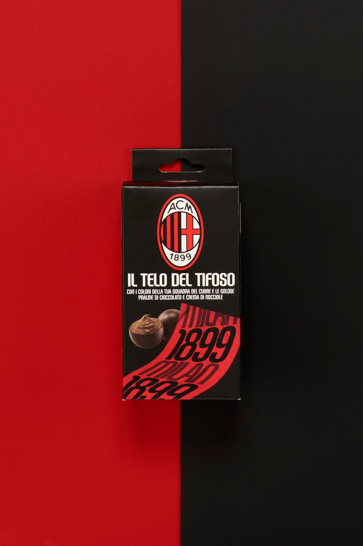 Confezione ufficiale AC Milan "Il Telo del Tifoso" posizionata al centro di uno sfondo diviso verticalmente nei colori iconici del club, rosso a sinistra e nero a destra. Sul fronte della scatola è raffigurato lo stemma AC Milan con la scritta "1899", accompagnato dalla dicitura "Il Telo del Tifoso". La confezione mostra anche una pralina di cioccolato ripiena alla crema di nocciole e un'anteprima del telo rosso personalizzato con grandi scritte nere e il numero 1899.