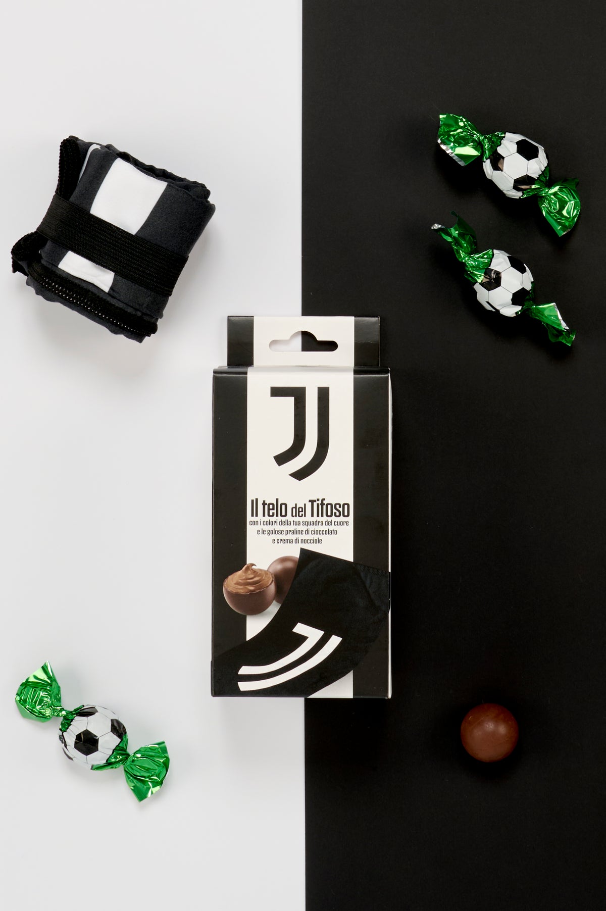 Confezione Juventus "Il Telo del Tifoso" fotografata dall'alto su uno sfondo metà bianco e metà nero. Attorno alla scatola sono disposti un telo nero piegato e fissato con una fascia elastica nera, tre praline incartate singolarmente con involucro verde metallizzato decorato a motivo pallone da calcio bianco e nero e una pralina di cioccolato senza confezione. L'immagine mette in evidenza tutti gli elementi del kit, creando un forte richiamo visivo ai colori caratteristici della squadra.