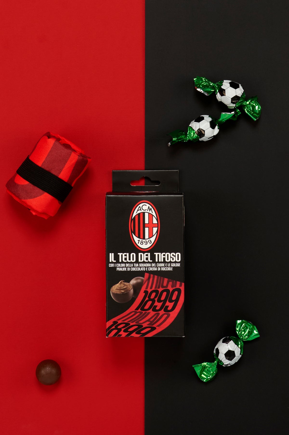 Confezione ufficiale AC Milan "Il Telo del Tifoso" fotografata dall'alto su uno sfondo metà rosso e metà nero. Attorno alla confezione sono disposti il telo piegato nei colori rossoneri, trattenuto da una fascia elastica nera, tre praline incartate singolarmente con confezione verde metallizzata decorata a motivo pallone da calcio bianco e nero e una pralina di cioccolato senza involucro.