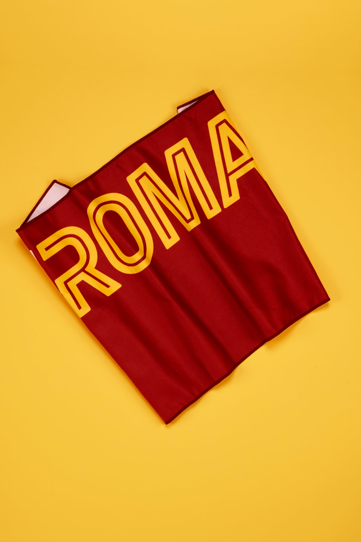 Telo ufficiale AS Roma disposto in diagonale su uno sfondo giallo uniforme. Il telo è realizzato in tessuto rosso intenso e presenta la grande scritta "ROMA" in giallo con bordo rosso, estesa per gran parte della superficie. Le leggere pieghe del tessuto ne evidenziano la morbidezza e la consistenza del materiale. L'inquadratura dall'alto valorizza il contrasto tra il rosso e il giallo, colori distintivi della squadra, e mette in risalto le dimensioni e il design del telo.