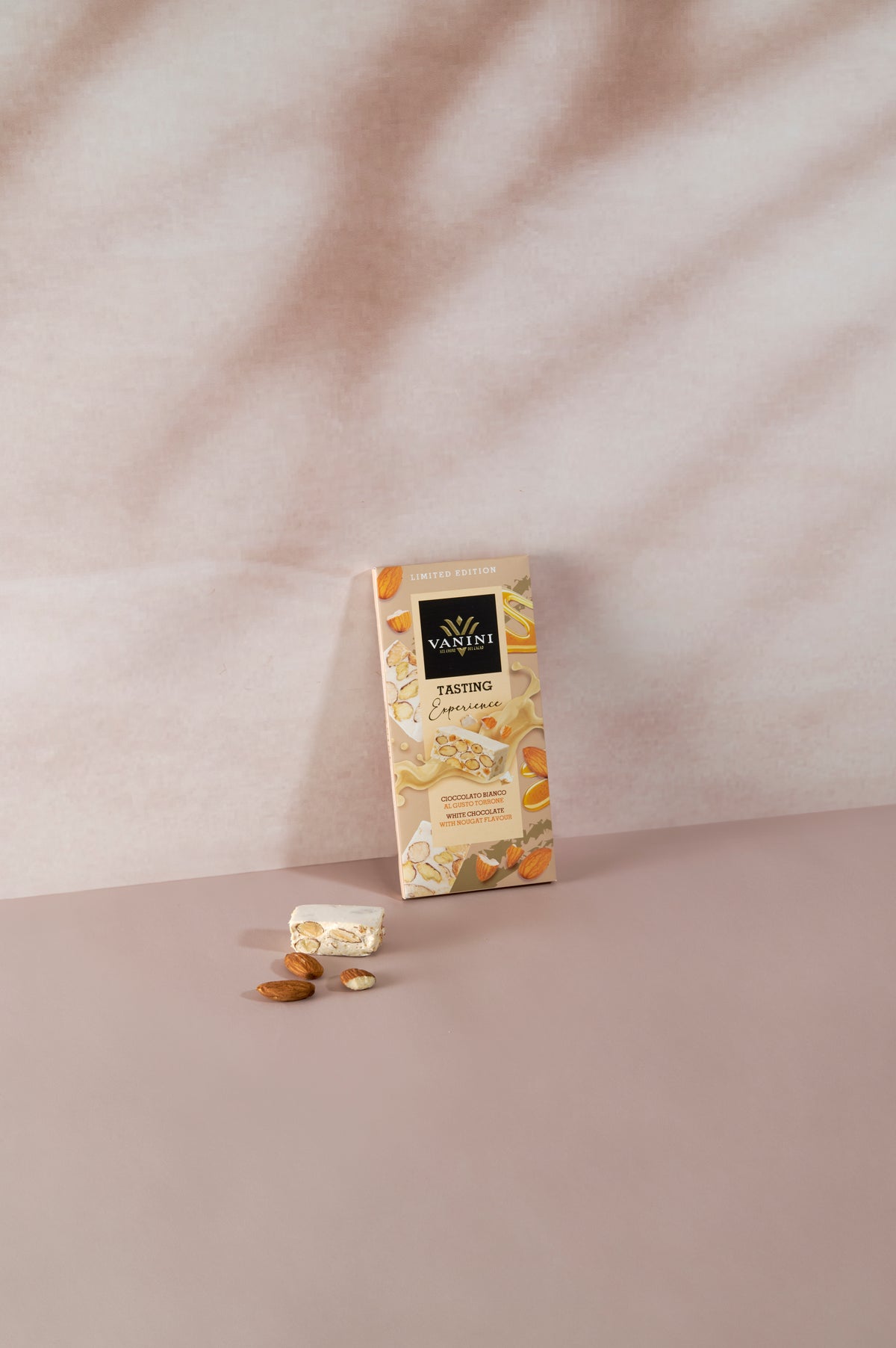 Tavoletta di cioccolato bianco Vanini Tasting Experience gusto torrone con mandorle, accompagnata da un pezzo di torrone e mandorle intere su sfondo dai toni rosa tenue.