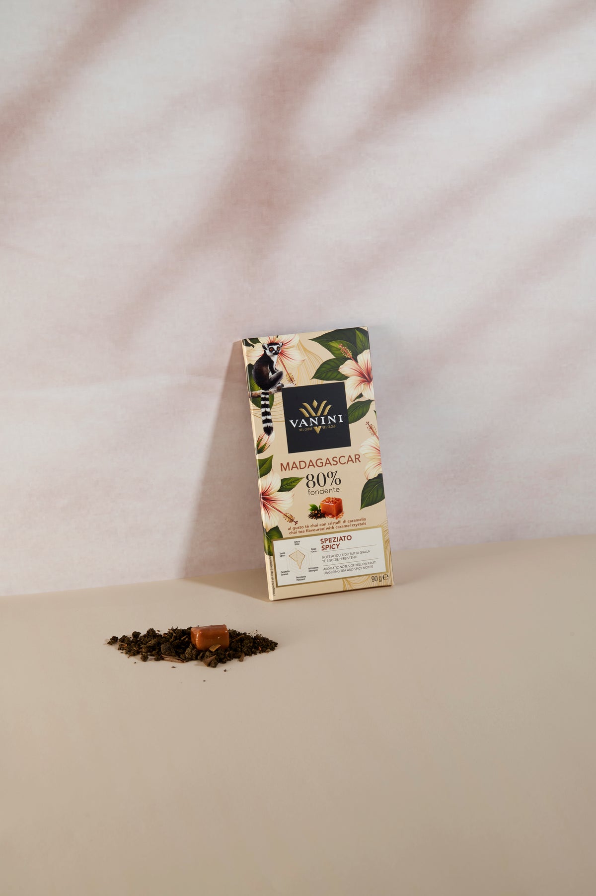 Tavoletta di cioccolato fondente Vanini Madagascar 80% con tè chai e cristalli di caramello, mostrata senza confezione. La tavoletta di cioccolato fondente è appoggiata verticalmente su uno sfondo beige chiaro; in primo piano si vedono foglie di tè chai sfuse e un cristallo di caramello che richiamano gli ingredienti della ricetta.