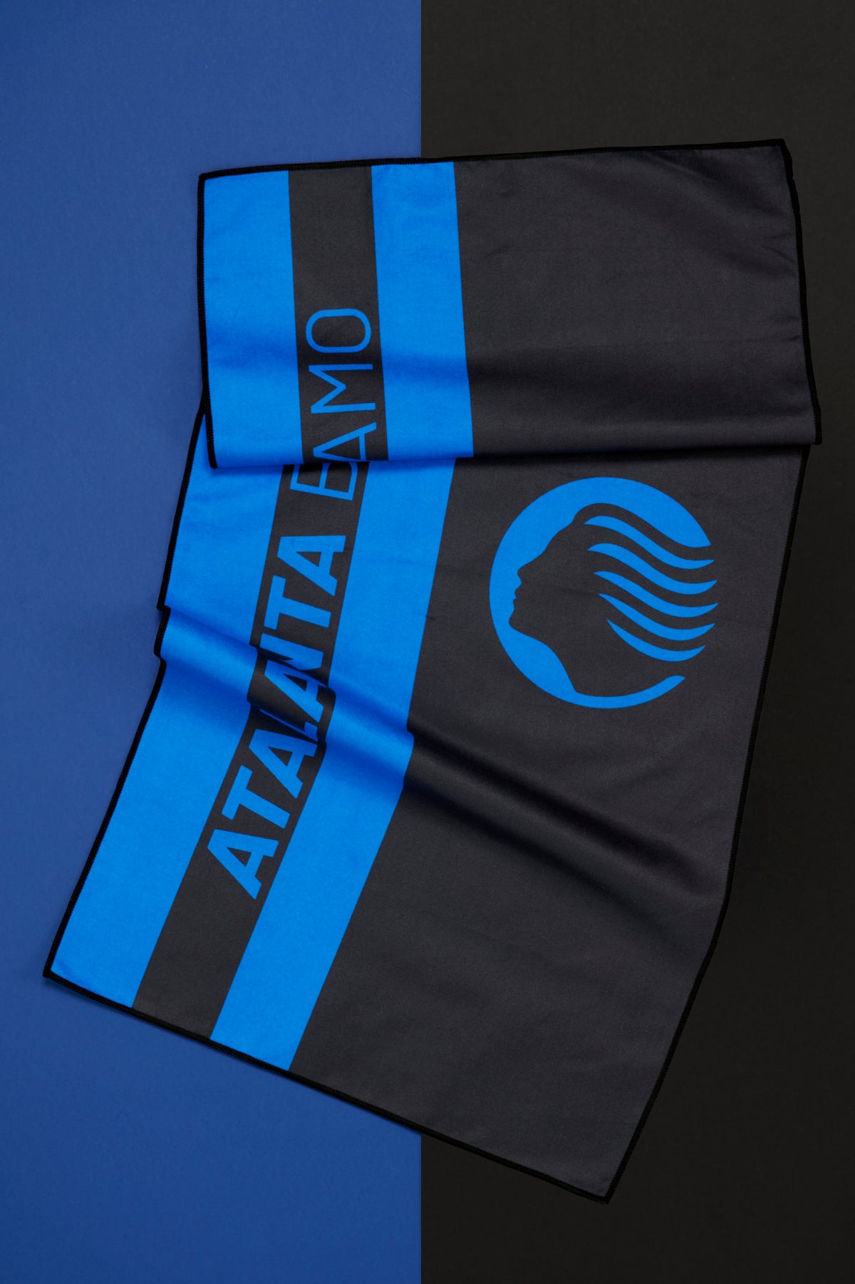 Telo Atalanta parzialmente aperto e disposto diagonalmente su uno sfondo diviso verticalmente nei colori blu e nero. Il tessuto presenta ampie bande verticali blu e nere e la scritta "ATALANTA BERGAMO" in blu lungo una fascia centrale. Nella parte inferiore è visibile il simbolo dell'Atalanta con il profilo stilizzato del volto caratterizzato da linee che richiamano una chioma al vento.
