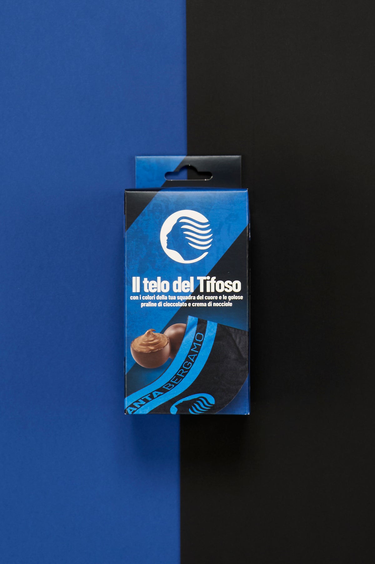 Confezione Atalanta "Il Telo del Tifoso" posizionata al centro di uno sfondo diviso verticalmente nei colori blu e nero. Sul fronte della scatola è visibile il profilo stilizzato del volto simbolo dell'Atalanta in bianco su sfondo blu, accompagnato dalla scritta "Il telo del Tifoso". Nella parte inferiore sono raffigurate praline di cioccolato ripiene alla crema di nocciole e un dettaglio del telo nero e blu con la scritta "Atalanta Bergamo". 