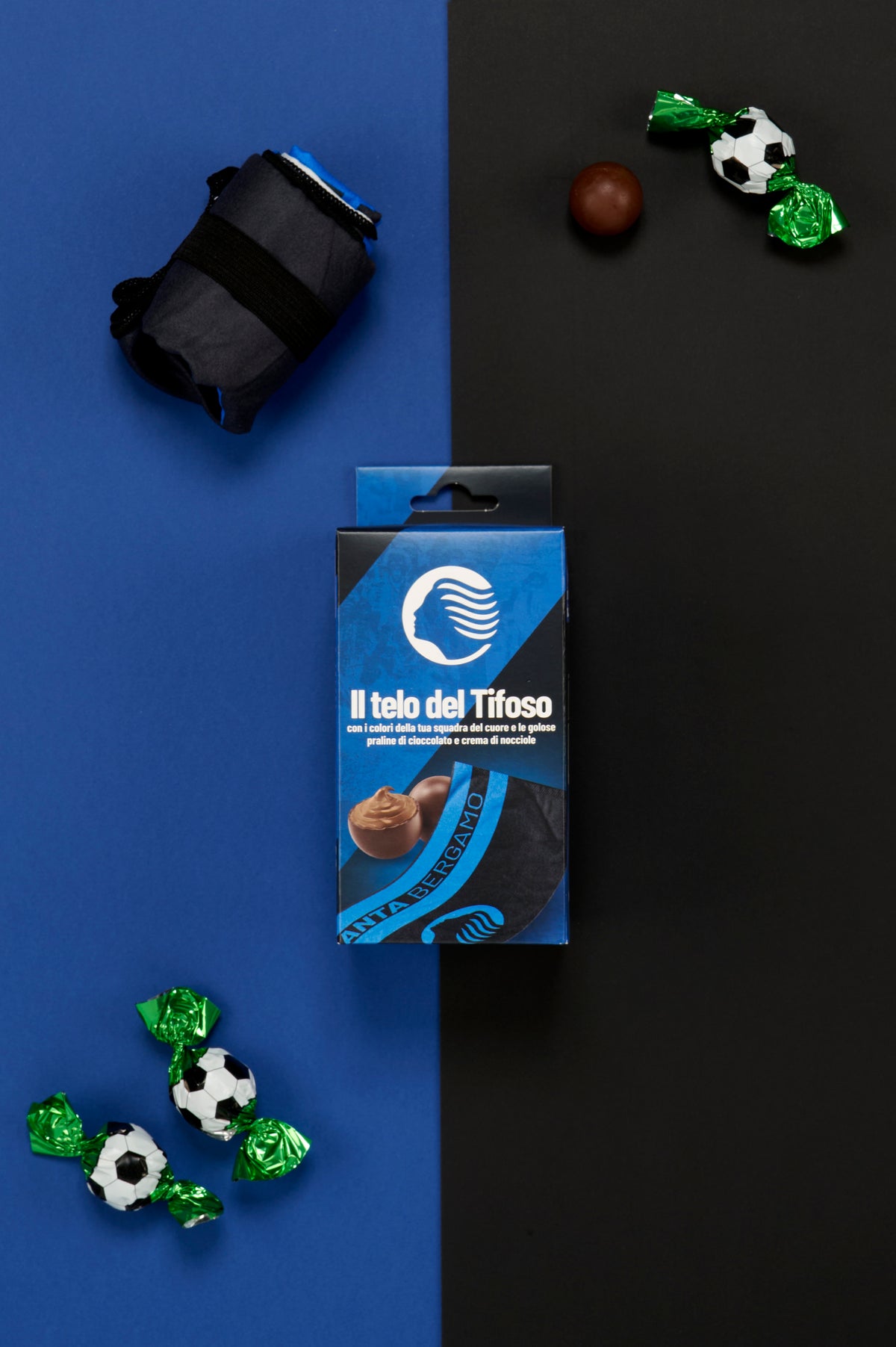Confezione Atalanta "Il Telo del Tifoso" fotografata dall'alto su uno sfondo metà blu e metà nero. Al centro dell'immagine è posizionata la scatola del prodotto, affiancata da un telo piegato nei colori della squadra e trattenuto da una fascia elastica nera. Attorno alla confezione sono disposte tre praline incartate singolarmente con involucro verde metallizzato decorato a motivo pallone da calcio bianco e nero e una pralina di cioccolato senza confezione. 