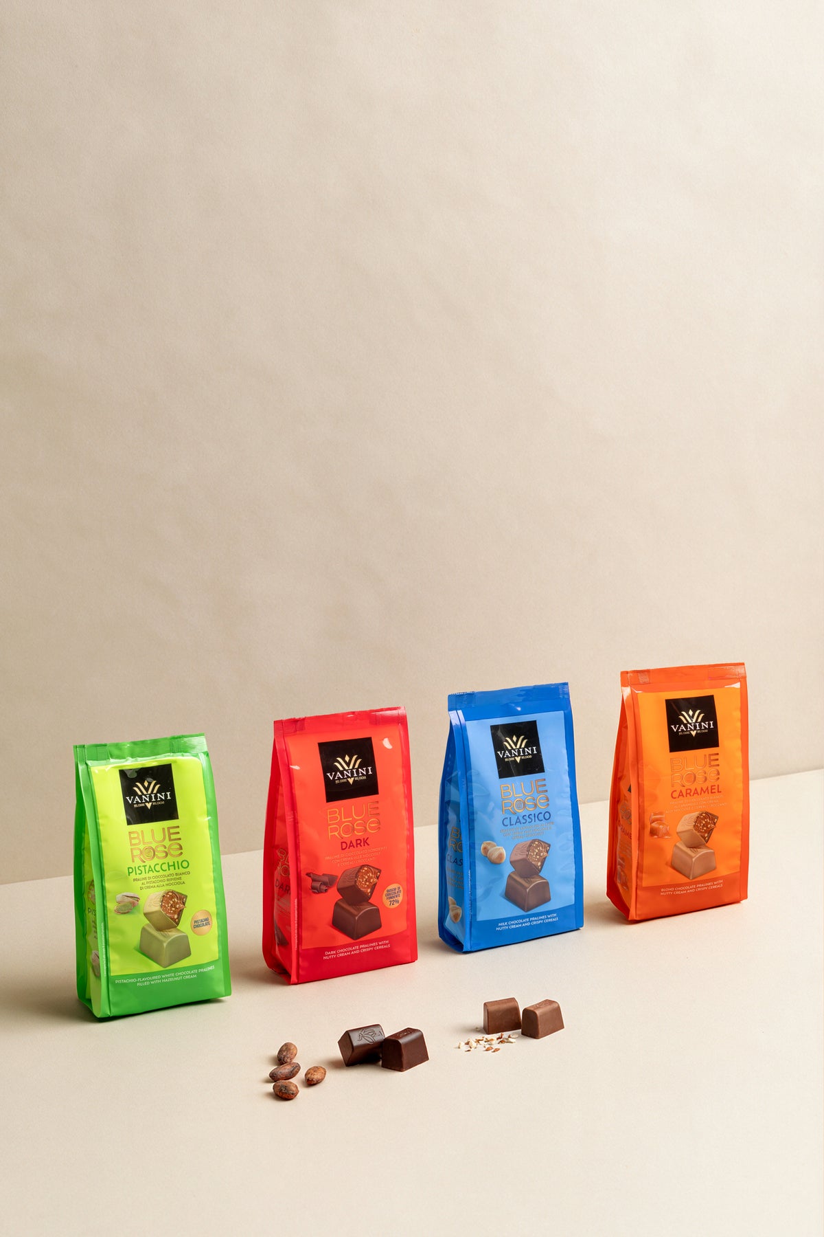 Confezione assortita della linea Vanini Blue Rose disposta su una superficie color avorio, davanti a uno sfondo neutro beige. Nell’immagine sono presenti quattro sacchetti di praline in piedi, allineati da sinistra a destra: Pistacchio in confezione verde brillante, Dark in confezione rosso acceso, Classico in confezione azzurra e Caramel in confezione arancione. Ogni confezione riporta il logo Vanini nella parte superiore e l’immagine della pralina corrispondente al gusto. 