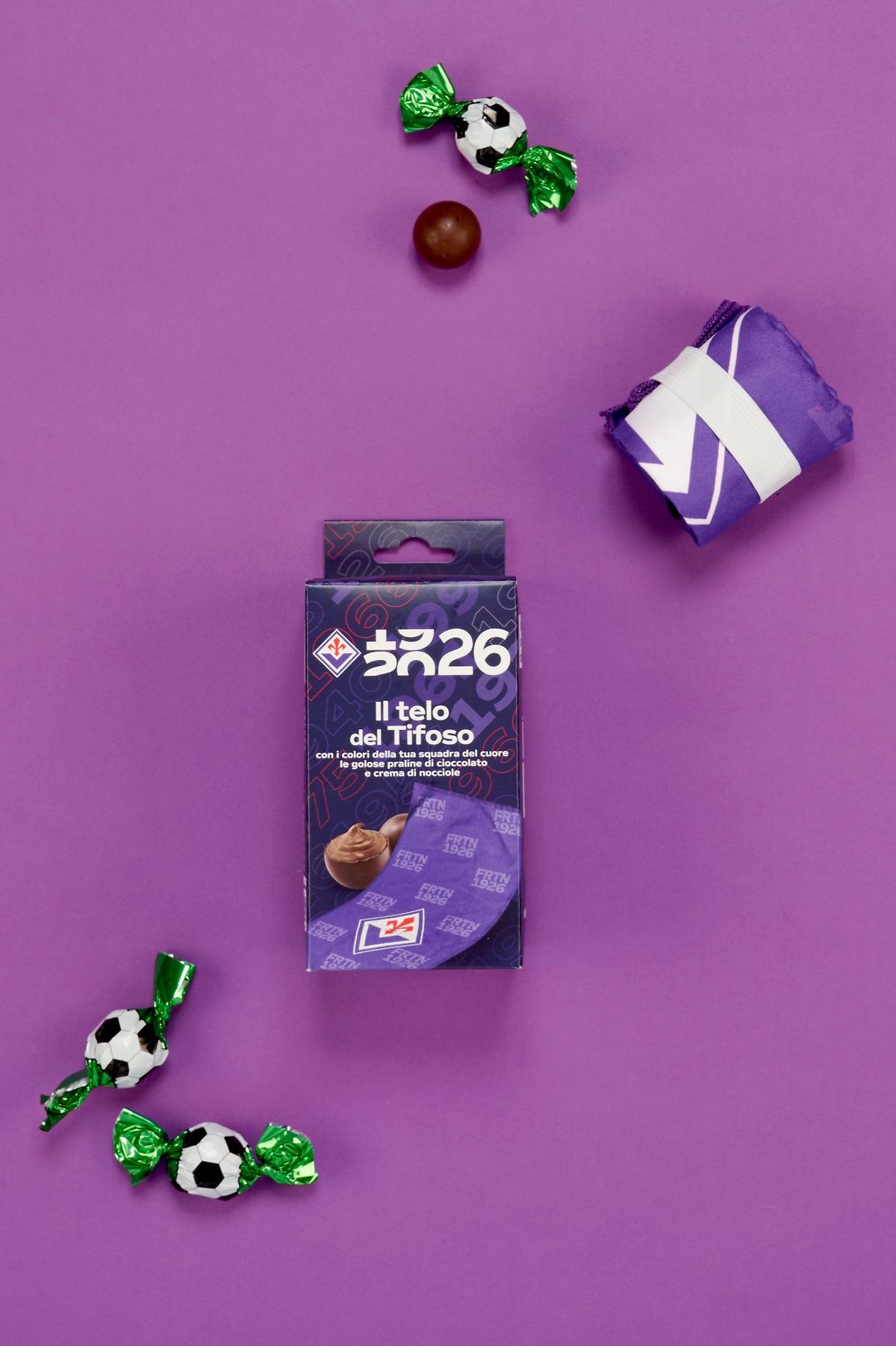 Confezione Fiorentina "Il Telo del Tifoso" fotografata dall'alto su uno sfondo viola. Accanto alla scatola è presente un telo piegato di colore viola, trattenuto da una fascia elastica bianca. Attorno al prodotto sono disposte tre praline incartate singolarmente con involucro verde metallizzato decorato a motivo pallone da calcio bianco e nero e una pralina di cioccolato senza confezione. L'immagine mostra tutti gli elementi del kit, evidenziando il richiamo ai colori e ai simboli della Fiorentina.