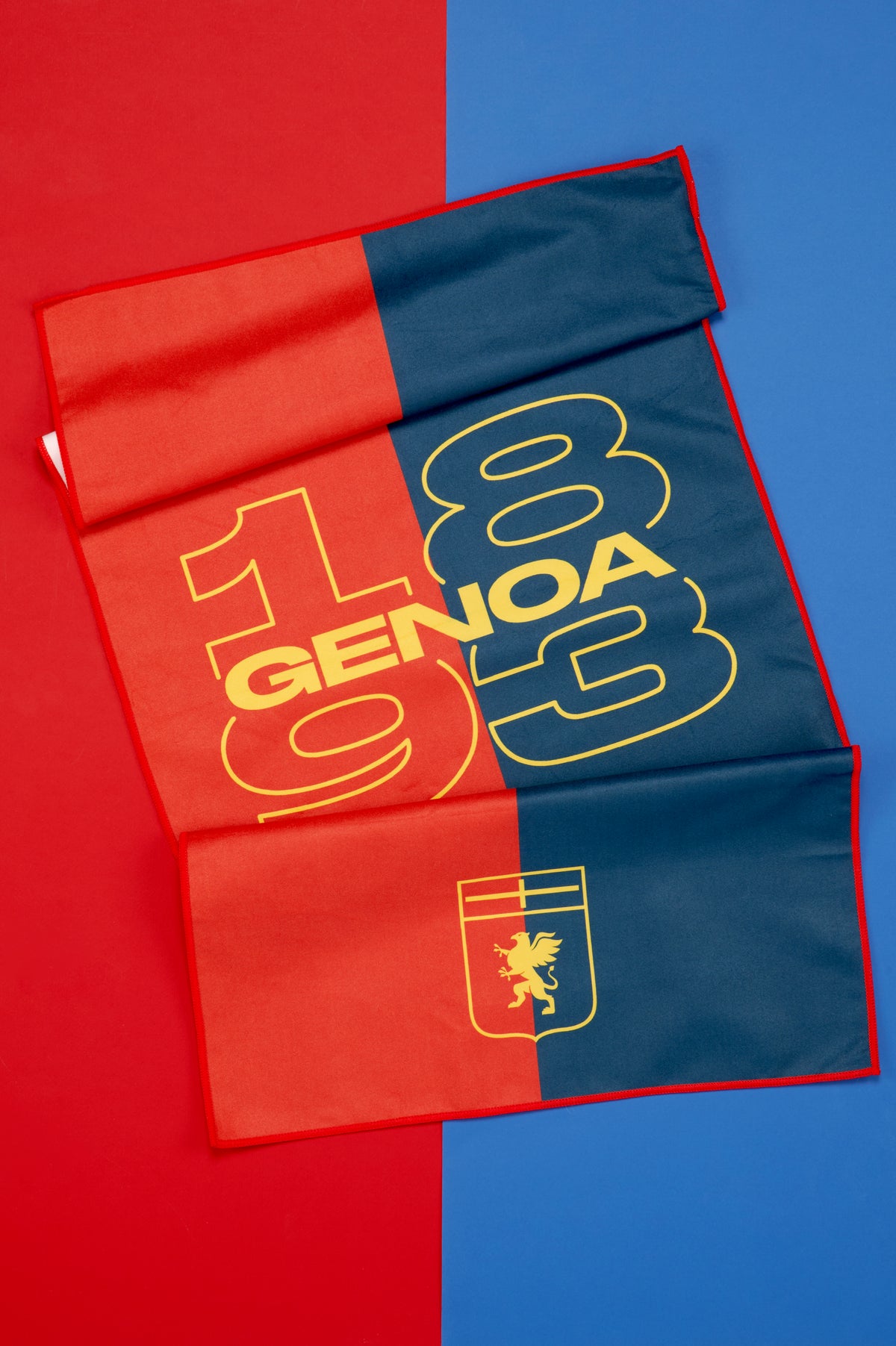 Telo Genoa parzialmente aperto e disposto su uno sfondo diviso verticalmente nei colori rosso e blu. Il tessuto presenta una grafica bicolore con ampi numeri "1893" e la scritta "GENOA" in giallo al centro della superficie. Nella parte inferiore è visibile lo stemma del Genoa con il grifone dorato su fondo rosso e blu. Le pieghe del tessuto mettono in risalto la morbidezza del materiale e le dimensioni del telo.