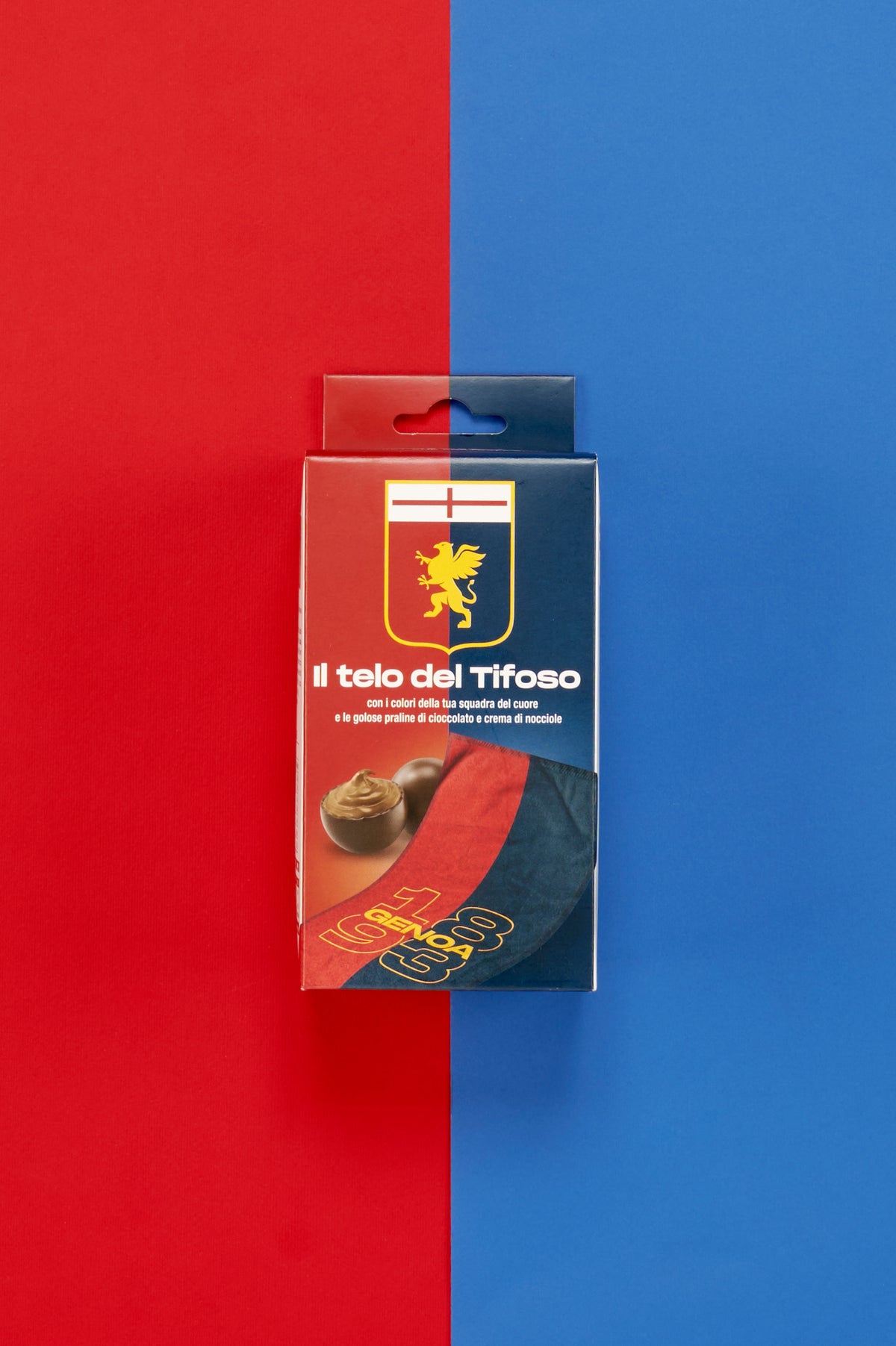 Confezione Genoa "Il Telo del Tifoso" posizionata al centro di uno sfondo diviso verticalmente nei colori rosso e blu. Sul fronte della scatola è visibile lo stemma del Genoa con il grifone dorato e la croce rossa nella parte superiore, accompagnato dalla scritta "Il telo del Tifoso". Nella parte inferiore sono raffigurate praline di cioccolato ripiene alla crema di nocciole e un dettaglio del telo personalizzato con grafica rosso e blu e la scritta "Genoa 1893" in giallo.