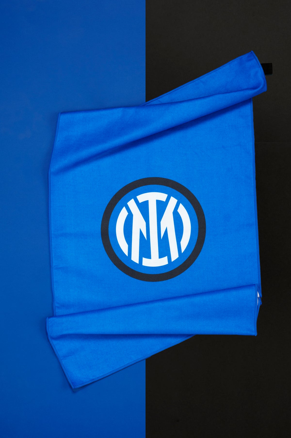 Telo Inter parzialmente aperto e disposto su uno sfondo diviso verticalmente nei colori blu e nero. Il tessuto blu presenta al centro il logo circolare dell'Inter in bianco racchiuso da un bordo nero, ben visibile grazie al forte contrasto cromatico. Le pieghe del tessuto mettono in risalto la morbidezza del materiale e le dimensioni del telo. La fotografia valorizza il design essenziale del prodotto e i colori distintivi associati alla squadra.