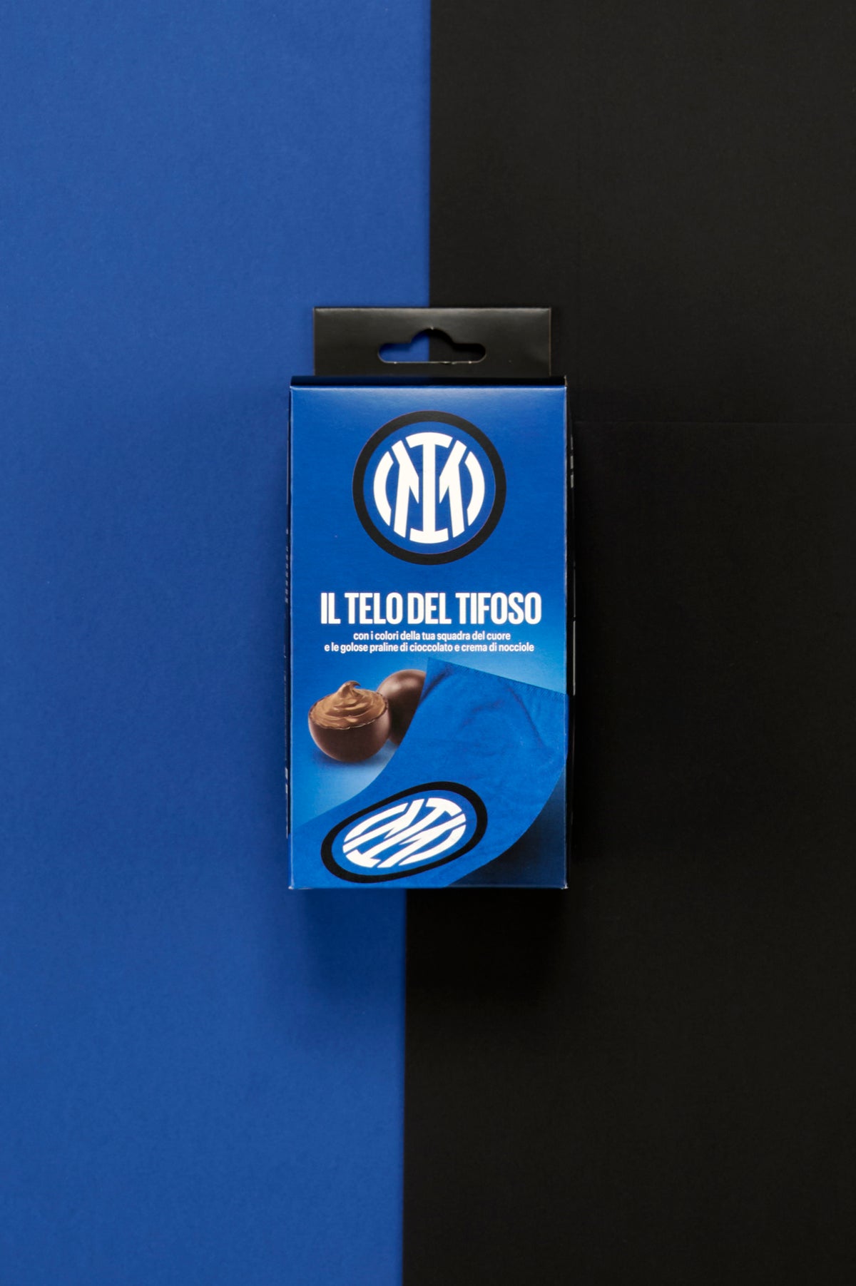 Confezione Inter "Il Telo del Tifoso" posizionata al centro di uno sfondo diviso verticalmente nei colori blu e nero. Sul fronte della scatola è visibile il logo circolare dell'Inter in bianco e nero su fondo blu, accompagnato dalla scritta "Il Telo del Tifoso". Nella parte inferiore sono raffigurate praline di cioccolato ripiene alla crema di nocciole e un dettaglio del telo blu personalizzato con il simbolo della squadra.