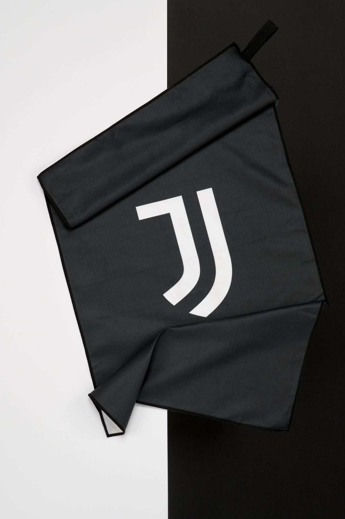 Telo Juventus parzialmente aperto e disposto diagonalmente su uno sfondo diviso verticalmente in bianco e nero. Il tessuto nero presenta al centro il logo stilizzato della Juventus in bianco, ben visibile grazie al forte contrasto cromatico. Alcune pieghe naturali del telo ne evidenziano la morbidezza e la texture del materiale. La fotografia valorizza il design minimalista del prodotto e l'identità visiva della squadra attraverso i suoi colori distintivi.