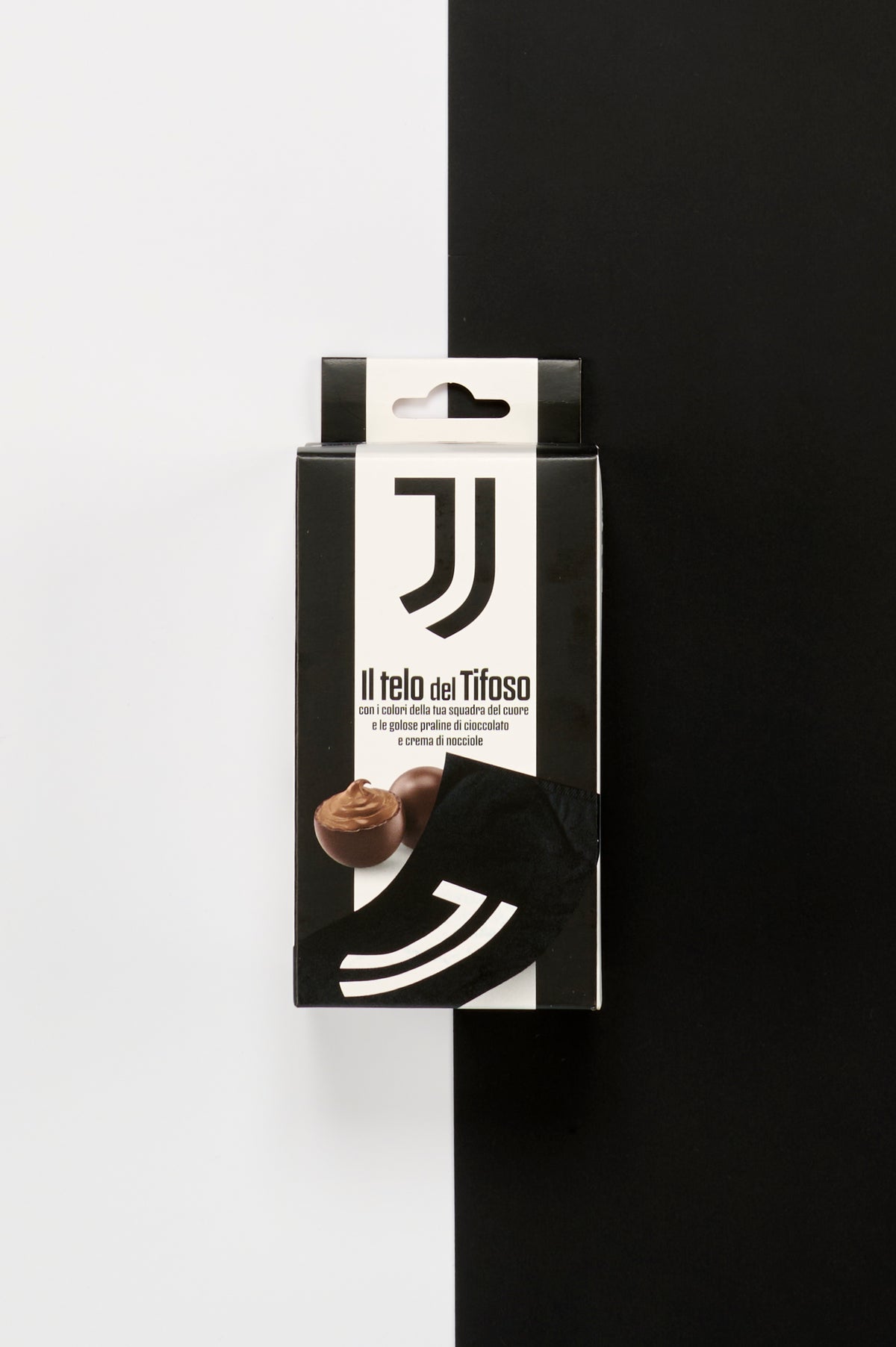 Confezione Juventus "Il Telo del Tifoso" posizionata al centro di uno sfondo diviso verticalmente in bianco e nero. Sul fronte della scatola è presente il logo stilizzato della Juventus in nero su fascia bianca, accompagnato dalla scritta "Il Telo del Tifoso". Nella parte inferiore sono raffigurate praline di cioccolato ripiene alla crema di nocciole e un dettaglio del telo nero personalizzato con il simbolo della squadra.