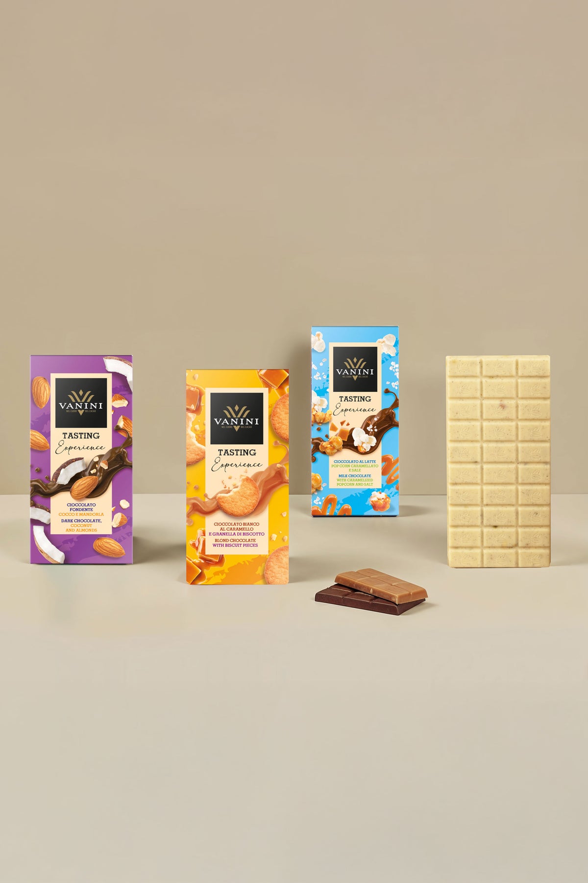 Selezione di tavolette Vanini Tasting Experience con esposizione di una tavoletta di cioccolato bianco aperta accanto alle confezioni della collezione. La composizione presenta gusti arricchiti da inclusioni selezionate come cocco, mandorle, granella di biscotto, popcorn caramellati, sale e pistacchio salato. Il design moderno e colorato delle confezioni è valorizzato da uno sfondo beige elegante che mette in risalto le diverse specialità di cioccolato.