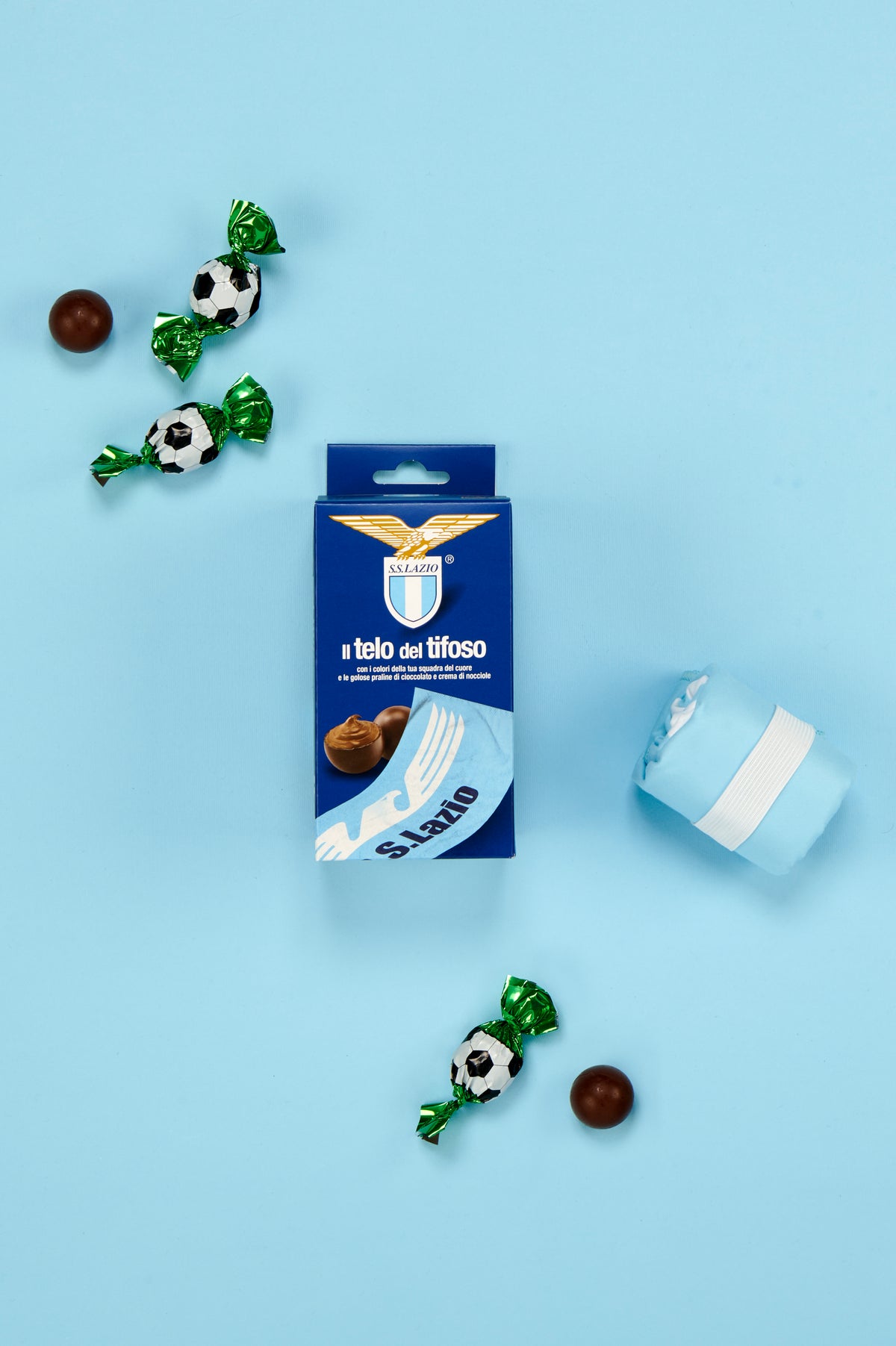 Confezione S.S. Lazio "Il telo del tifoso" fotografata dall'alto su sfondo azzurro. Al centro è posizionata la scatola blu con il simbolo dell'aquila e lo stemma della squadra, affiancata da un telo celeste piegato e trattenuto da una fascia elastica bianca. Attorno alla confezione sono disposte tre praline incartate singolarmente con involucro verde metallizzato decorato a motivo pallone da calcio bianco e nero, insieme a due praline di cioccolato prive di confezione.