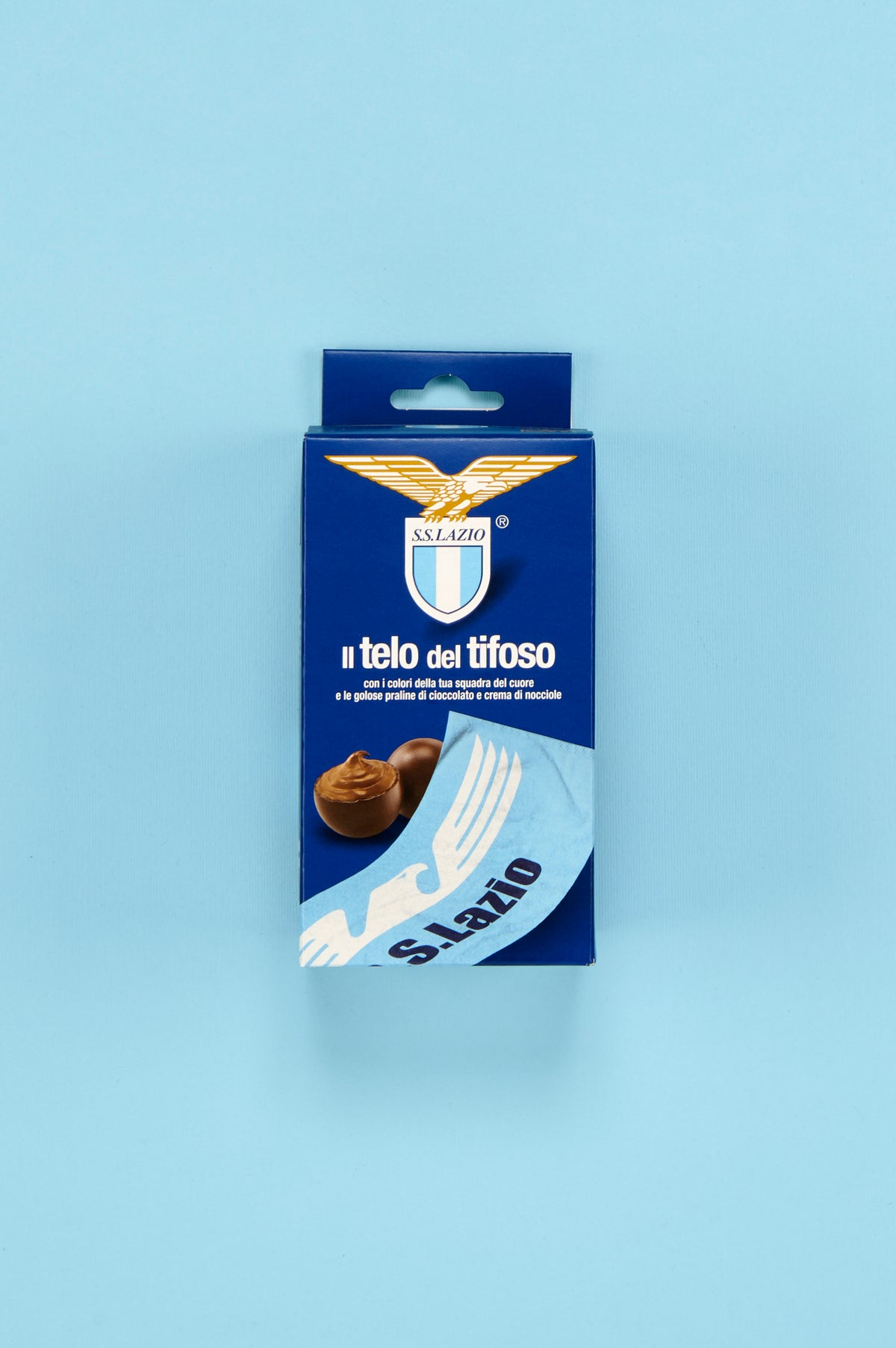 Confezione S.S. Lazio "Il telo del tifoso" posizionata al centro di uno sfondo azzurro uniforme. Sul fronte della scatola è visibile il simbolo dell'aquila dorata con lo stemma della squadra, la scritta "Il telo del tifoso" e l'immagine di praline di cioccolato ripiene alla crema di nocciole. Nella parte inferiore è raffigurato un dettaglio del telo celeste personalizzato con aquila stilizzata bianca e scritta "S.S. Lazio". L'inquadratura frontale valorizza il packaging e i colori distintivi del club.