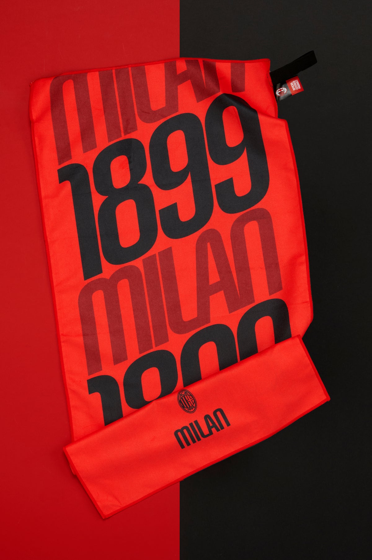 Telo ufficiale AC Milan disteso su uno sfondo bicolore rosso e nero. Il tessuto rosso presenta un design grafico d'impatto con la scritta "MILAN" e il numero "1899" ripetuti in grandi caratteri neri che occupano gran parte della superficie. Nella parte inferiore è visibile il logo AC Milan accompagnato dalla scritta "MILAN". Le leggere pieghe del tessuto evidenziano la morbidezza del materiale e le dimensioni del telo, mentre il contrasto tra rosso e nero richiama immediatamente i colori storici del club.