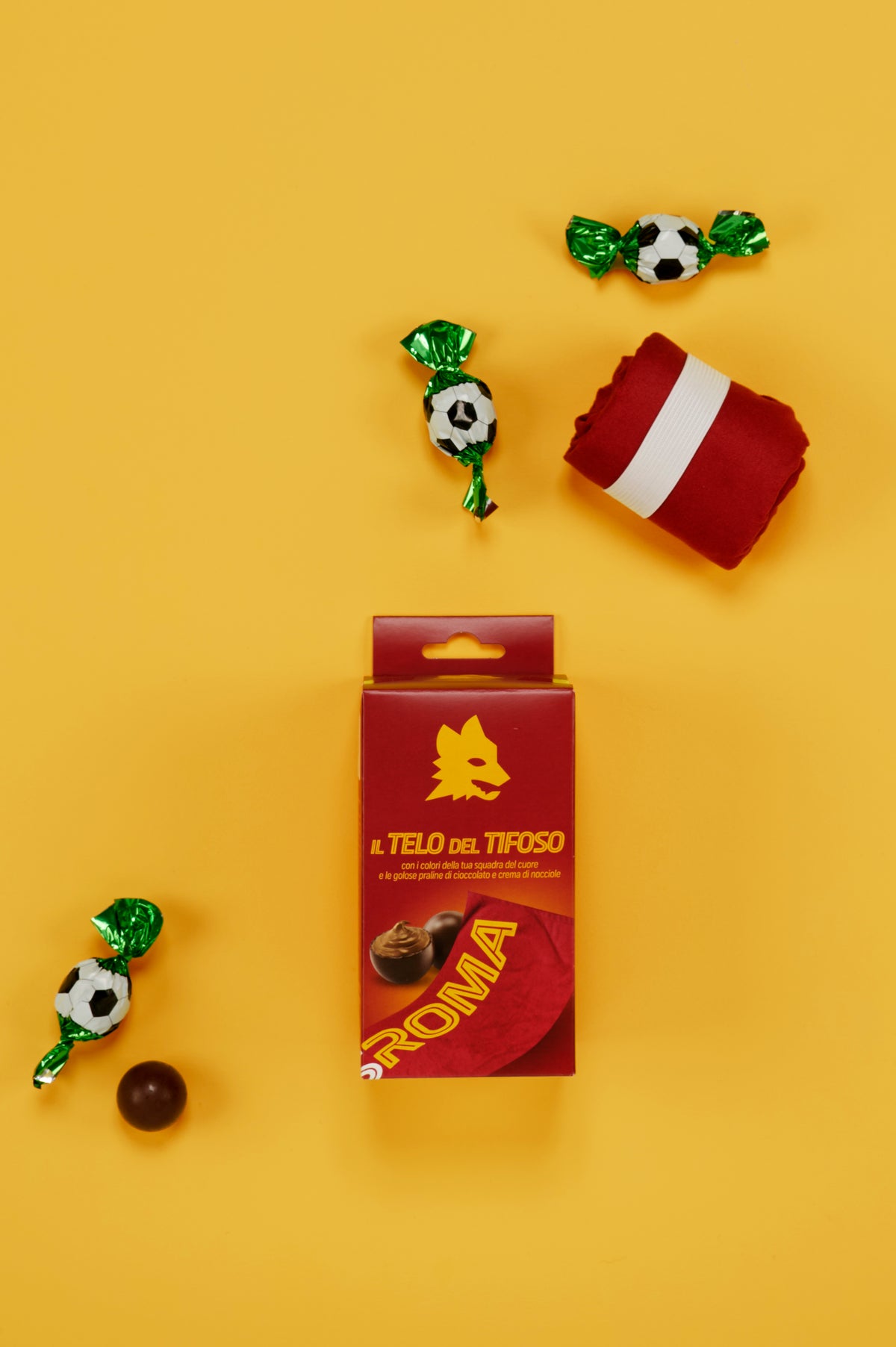 Confezione ufficiale AS Roma "Il Telo del Tifoso" fotografata dall'alto su sfondo giallo uniforme. Al centro dell'immagine è posizionata la scatola nei colori sociali giallorossi, decorata con il simbolo del lupo stilizzato e l'immagine del telo personalizzato AS Roma. Attorno alla confezione sono disposte tre praline di cioccolato incartate singolarmente con involucro verde metallizzato e fantasia pallone da calcio bianco e nero, una pralina di cioccolato senza confezione e il telo piegato di colore rosso.