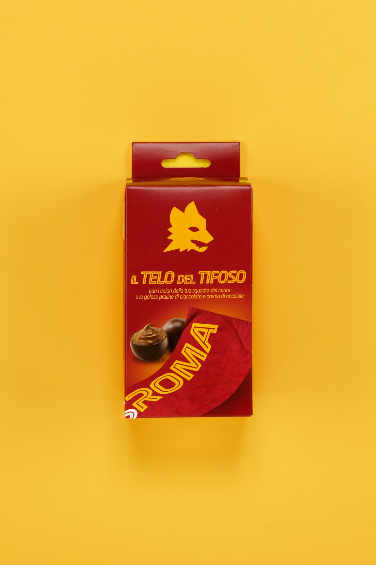 Confezione ufficiale AS Roma "Il Telo del Tifoso" posizionata al centro di uno sfondo giallo uniforme. Sul fronte della scatola è visibile il logo del lupo stilizzato in giallo su fondo rosso, accompagnato dalla scritta "Il Telo del Tifoso". La confezione mostra inoltre l'immagine di praline di cioccolato ripiene alla crema di nocciole e una porzione del telo rosso personalizzato con la scritta "ROMA". 
