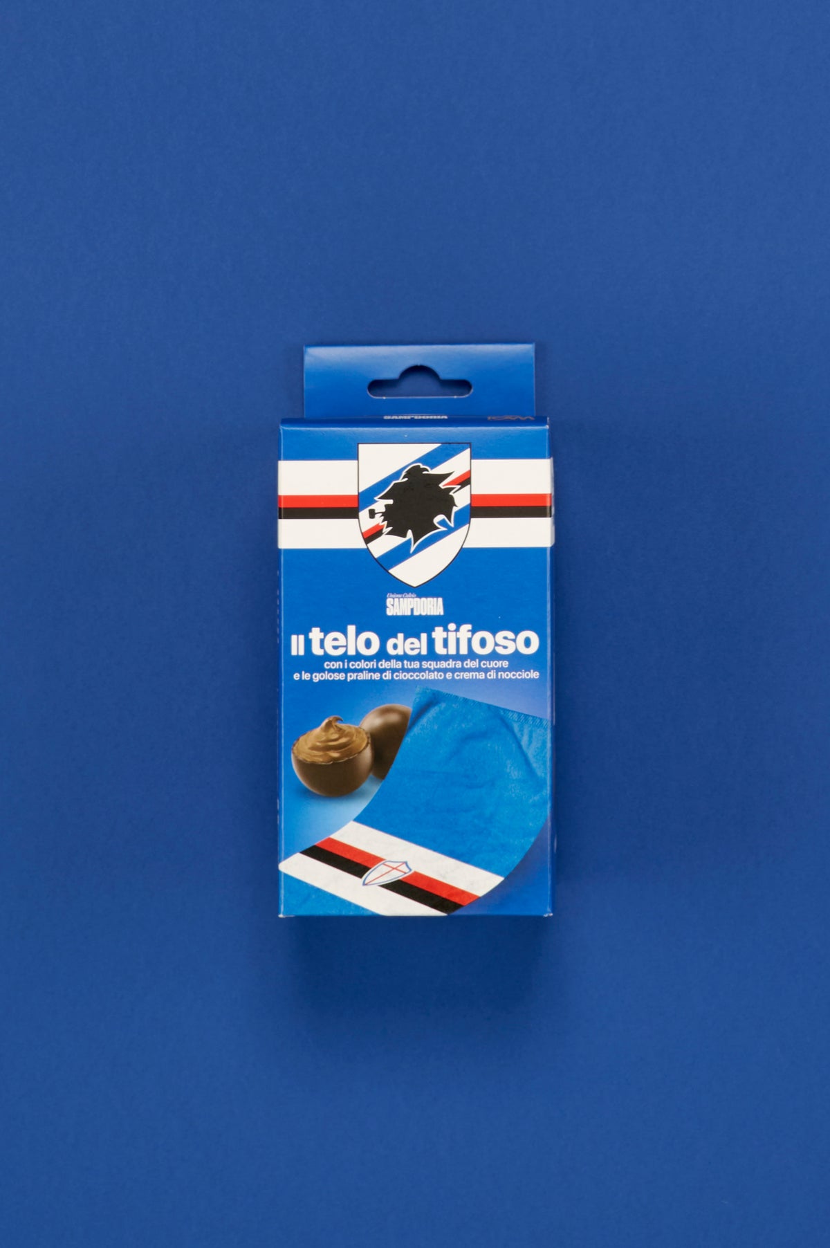 Confezione ufficiale UC Sampdoria "Il telo del tifoso" fotografata frontalmente e posizionata al centro di uno sfondo blu uniforme. Sul fronte della scatola è visibile lo stemma della Sampdoria con il profilo del marinaio e le iconiche bande orizzontali bianca, blu, rossa e nera. La confezione riporta la scritta "Il telo del tifoso" e l'immagine del telo azzurro decorato con i colori sociali del club. Sono inoltre raffigurate praline di cioccolato ripiene alla crema di nocciole.