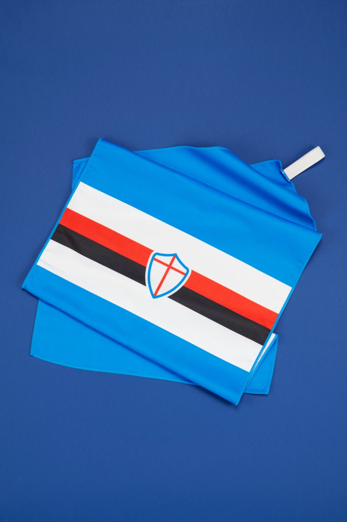 Telo ufficiale UC Sampdoria parzialmente aperto e disposto su uno sfondo blu uniforme. Il telo è realizzato in tonalità azzurra e presenta al centro le caratteristiche bande orizzontali bianca, rossa e nera della Sampdoria, arricchite dallo scudetto con la croce rossa bordata di blu. Il tessuto è ripiegato in alcuni punti per evidenziare morbidezza, dimensioni e finitura del materiale. La fotografia valorizza l'identità visiva del club attraverso i suoi colori distintivi e i simboli più rappresentativi.
