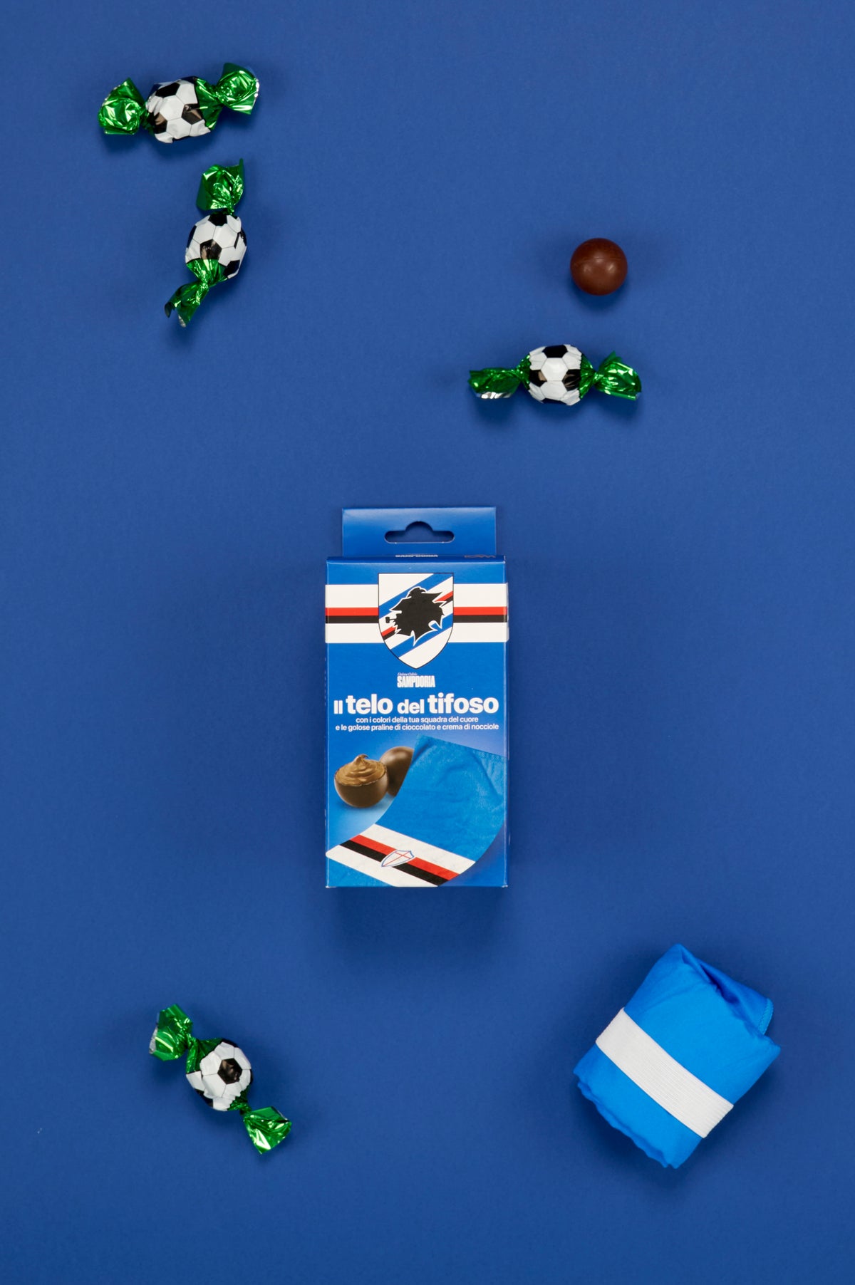 Confezione UC Sampdoria "Il telo del tifoso" disposta al centro di una composizione fotografata dall'alto su sfondo blu. Attorno alla confezione sono posizionate tre praline incartate singolarmente con involucro verde metallizzato e fantasia pallone da calcio bianco e nero, una pralina di cioccolato senza confezione e il telo piegato nei colori della squadra, fissato con una fascia elastica bianca. La disposizione degli elementi richiama il mondo del tifo calcistico e mette in risalto il contenuto del kit.
