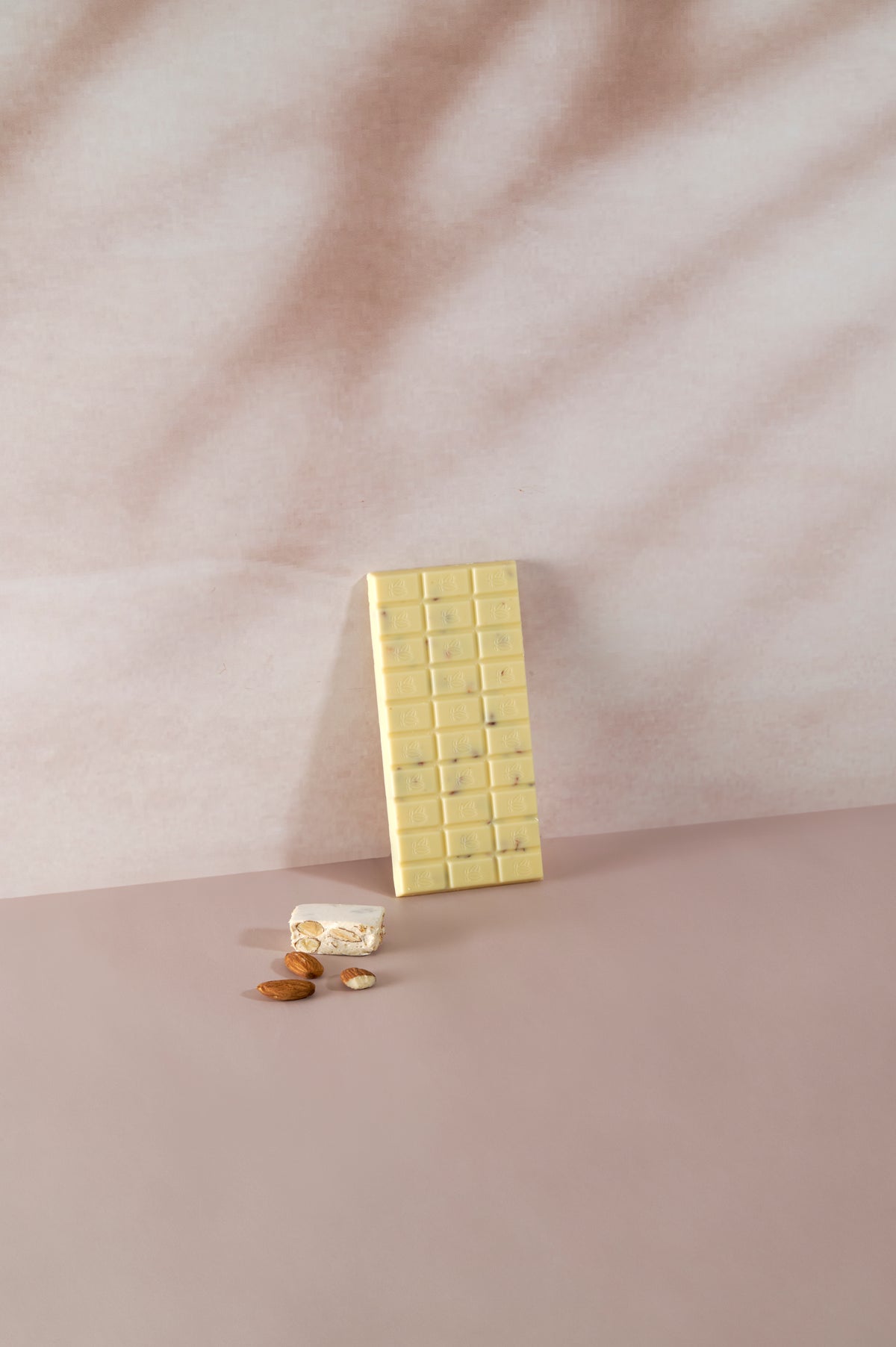 Tavoletta di cioccolato bianco Vanini Tasting Experience gusto torrone con inclusioni di mandorle, affiancata da un pezzo di torrone tradizionale e mandorle intere su sfondo rosa delicato.