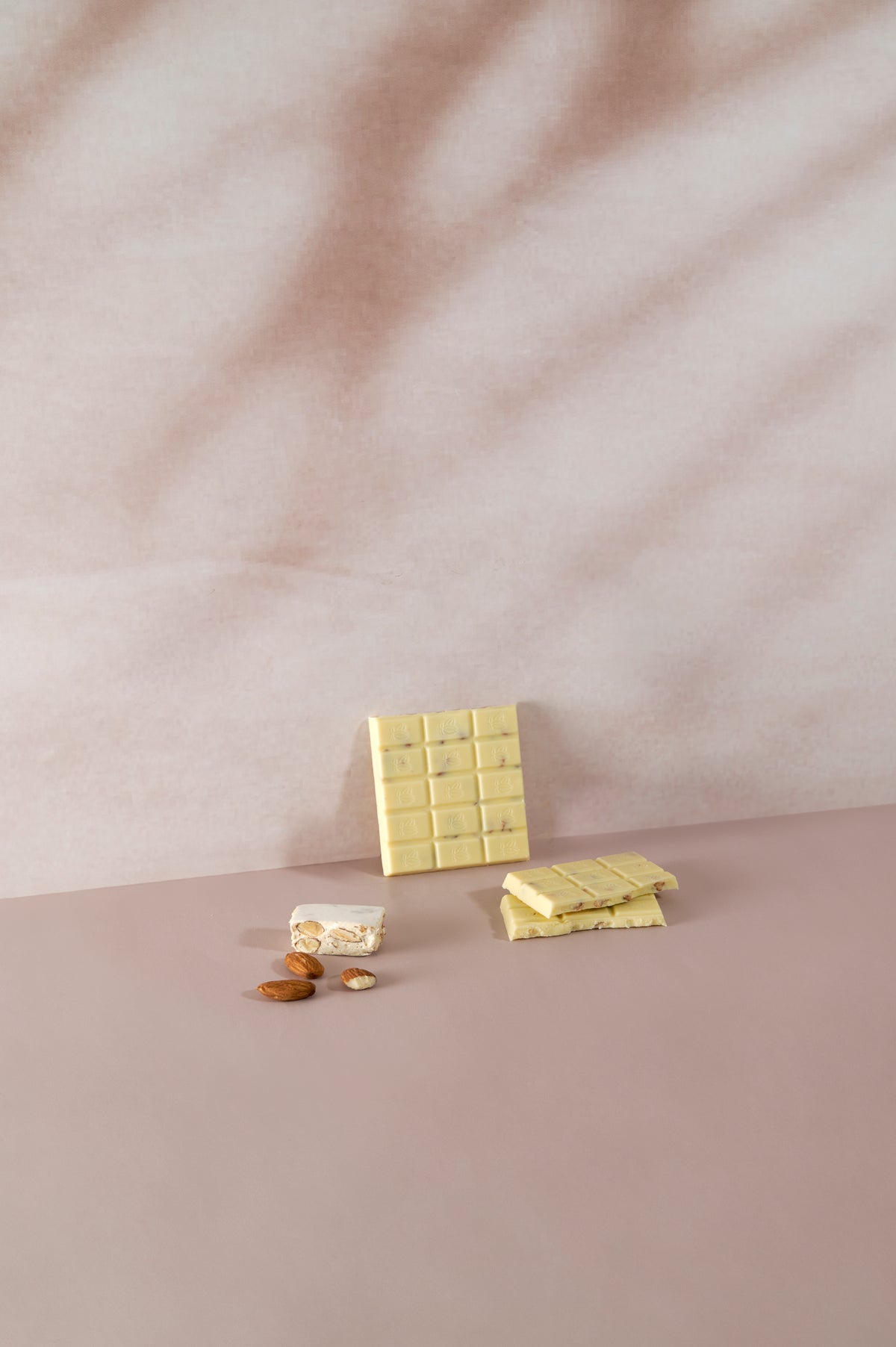 Tavoletta di cioccolato bianco Vanini Tasting Experience gusto torrone divisa a pezzi, con inclusioni di mandorle, affiancata da un pezzo di torrone tradizionale e mandorle intere su sfondo rosa delicato.