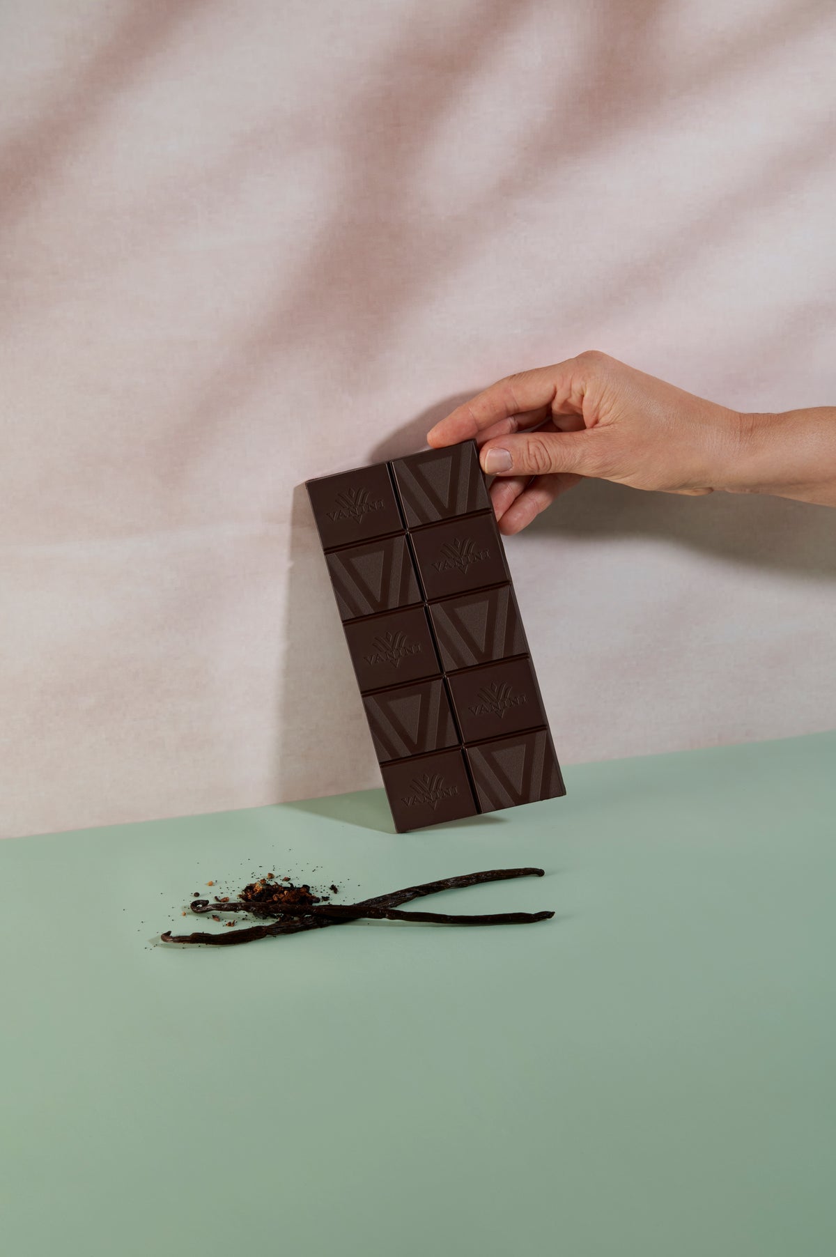 Mano che solleva una tavoletta di cioccolato fondente Vanini Giamaica 80% con note di vaniglia, ambientata in un set fotografico elegante con baccelli di vaniglia e cacao.