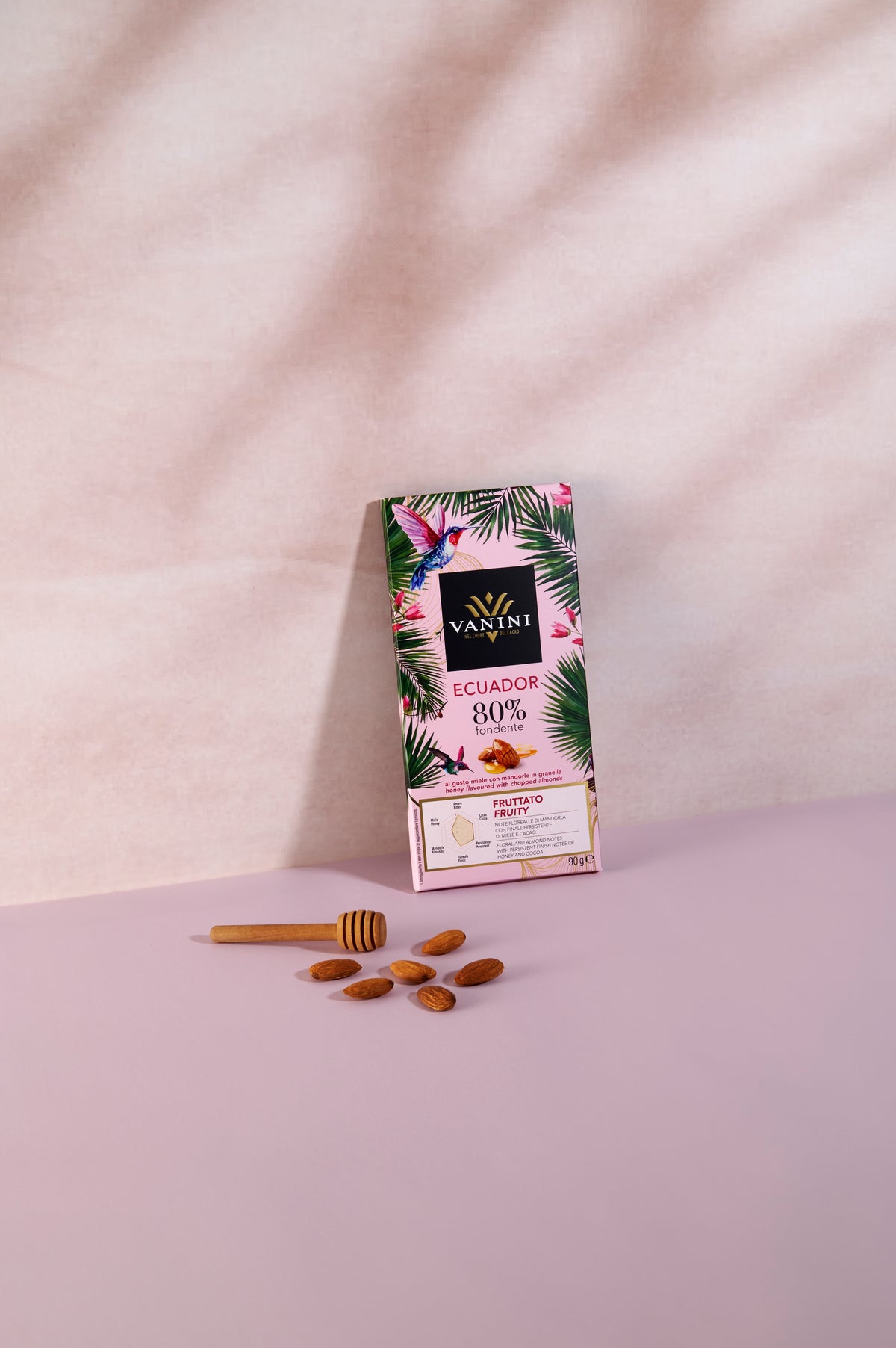 Tavoletta Vanini Ecuador 80% fondente con miele e mandorle in granella, confezione rosa decorata con foglie tropicali e colibrì colorati. Il prodotto è fotografato in verticale su una superficie rosa pastello, accompagnato da un cucchiaino spargimiele in legno e mandorle intere che richiamano gli ingredienti della ricetta. Lo sfondo chiaro con ombre delicate valorizza il carattere elegante e raffinato della tavoletta Monorigine Ecuador.