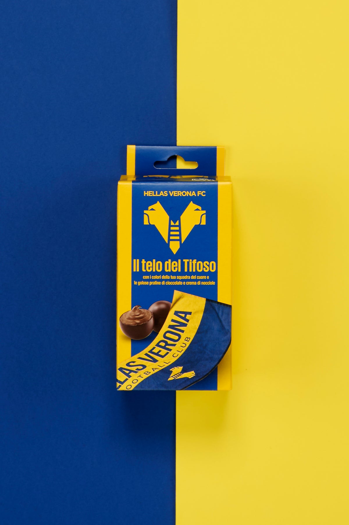 Confezione ufficiale Hellas Verona FC "Il telo del Tifoso" posizionata al centro di uno sfondo diviso verticalmente nei colori della squadra, blu a sinistra e giallo a destra. La scatola presenta il logo del club in giallo su fondo blu, la scritta "Il telo del Tifoso" e l'immagine di un telo gialloblù personalizzato con la scritta "Hellas Verona Football Club". Sul fronte sono raffigurate anche praline di cioccolato ripiene alla nocciola.