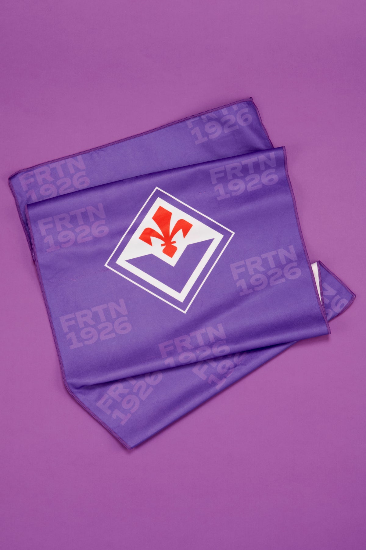 Telo Fiorentina parzialmente aperto e disposto su uno sfondo viola uniforme. Il tessuto presenta al centro il logo della squadra con il giglio rosso all'interno di un rombo bianco bordato di viola. Sulla superficie del telo sono presenti motivi tono su tono con la scritta "FRTN 1926" ripetuta in più punti. Le pieghe del tessuto ne evidenziano la morbidezza e la consistenza, mentre il colore viola dominante richiama immediatamente l'identità visiva della Fiorentina.
