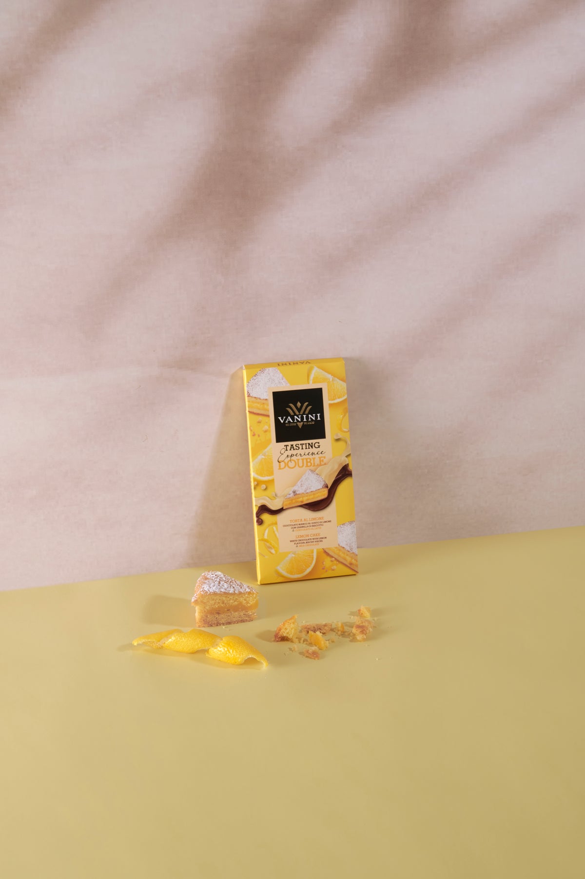 Confezione Vanini Tasting Experience Double Torta al Limone su sfondo dai toni gialli, affiancata da una fetta di torta al limone, scorza di limone e briciole di dolce che richiamano gli ingredienti della tavoletta.