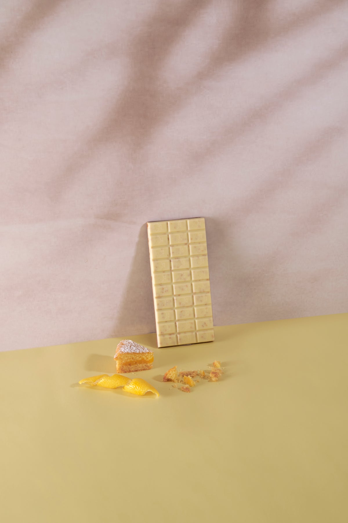 una tavoletta di cioccolato bianco scartata Vanini Tasting Experience Double Torta al Limone. Sul piano sottostante sono presenti una fetta di torta al limone, scorza di limone e briciole di dolce su sfondo giallo tenue.