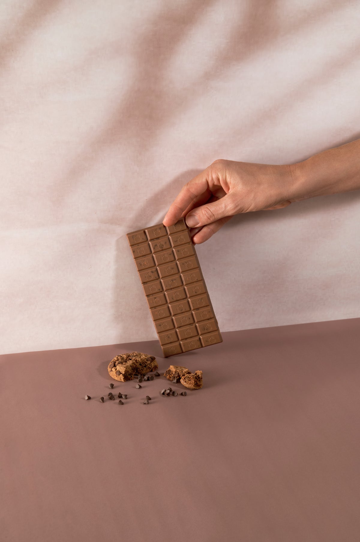 Mano che solleva una tavoletta di cioccolato al latte Vanini con cookies e gocce di cioccolato, su sfondo dai toni rosa e beige con biscotti sbriciolati in primo piano.