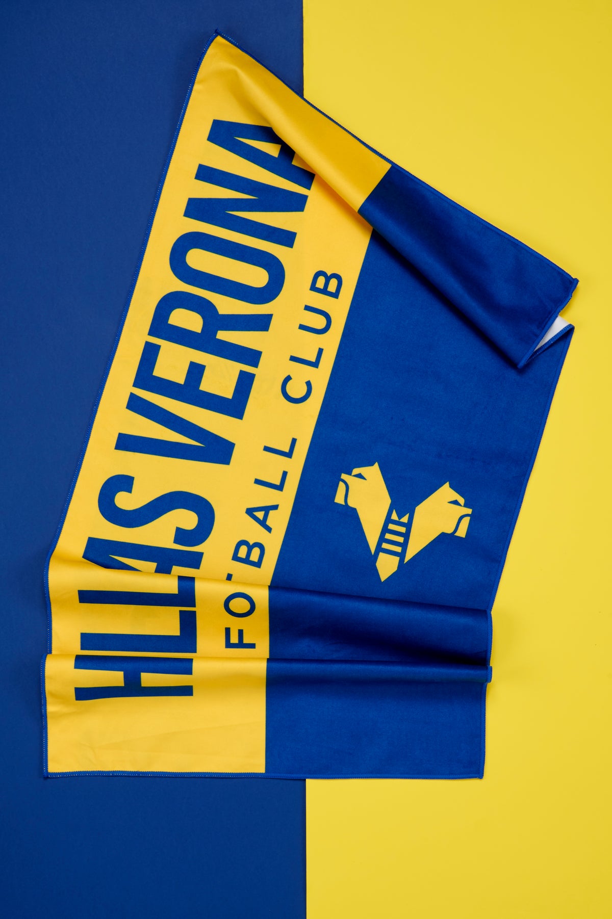 Telo ufficiale Hellas Verona FC aperto e disposto diagonalmente su uno sfondo bicolore blu e giallo. Il telo presenta un design a blocchi di colore con l'ampia scritta "HELLAS VERONA FOOTBALL CLUB" in blu su fascia gialla e il logo stilizzato del club in giallo su fondo blu. Il tessuto è leggermente piegato per mostrare il drappeggio e la consistenza del materiale.