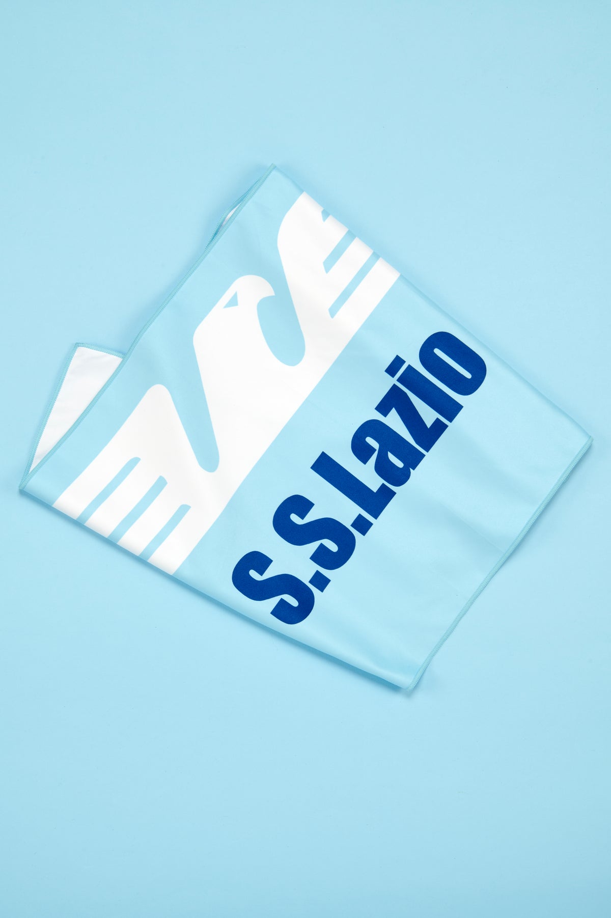 Telo S.S. Lazio disposto diagonalmente su uno sfondo azzurro uniforme. Il tessuto, di colore celeste, presenta una grande aquila stilizzata bianca che occupa gran parte della superficie e la scritta "S.S. Lazio" in blu scuro. Il telo è leggermente ripiegato per evidenziare la consistenza del materiale e le dimensioni del prodotto. La composizione minimalista richiama i colori tradizionali della squadra e mette in risalto il design grafico del telo.