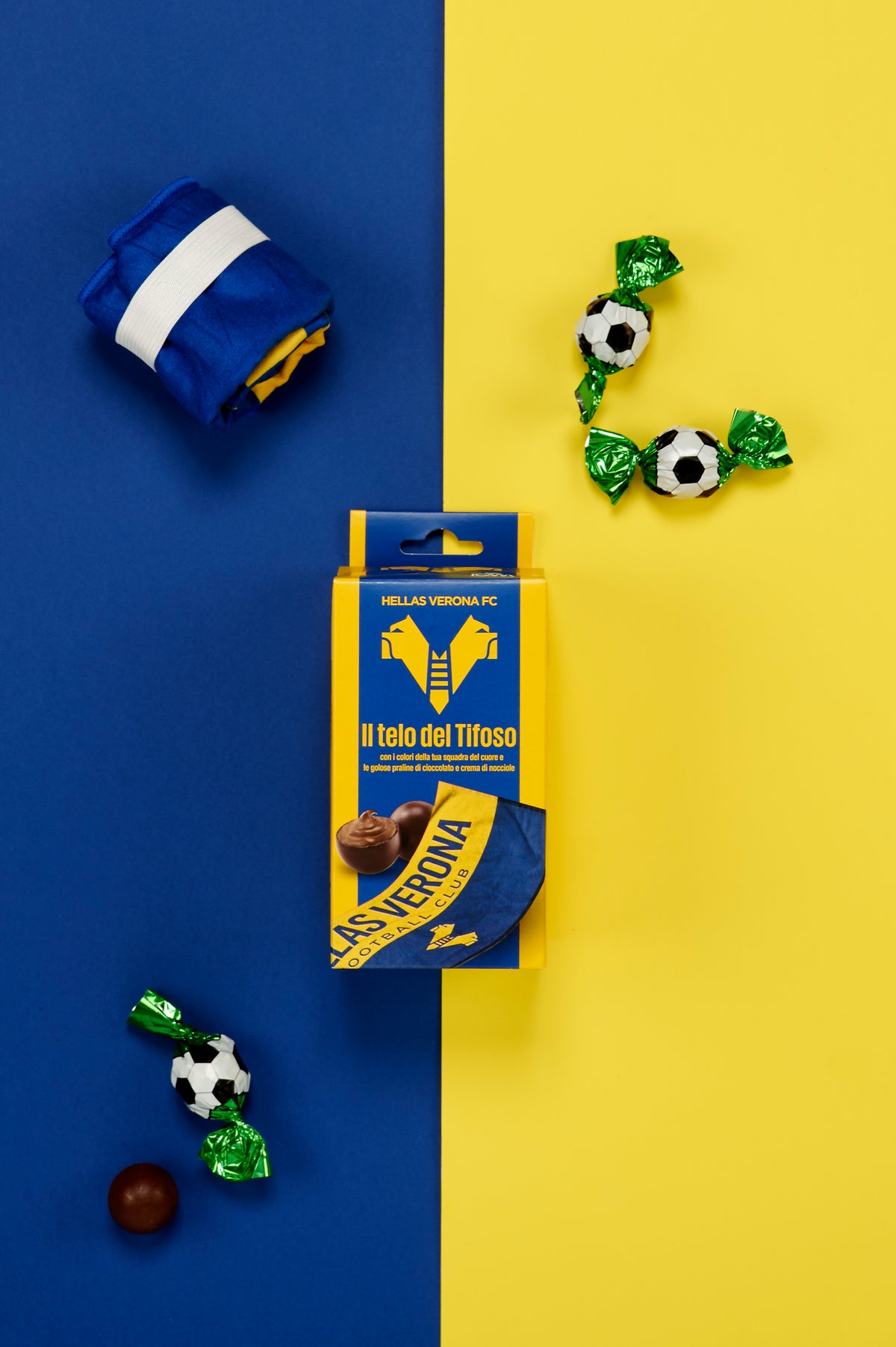 Confezione Hellas Verona FC "Il telo del Tifoso" fotografata dall'alto su uno sfondo metà blu e metà giallo. Accanto alla confezione sono disposti il telo piegato nei colori ufficiali del club, trattenuto da una fascia bianca, e tre praline di cioccolato incartate singolarmente in carta verde brillante con fantasia pallone da calcio bianco e nero. Una pralina è mostrata senza involucro, mettendo in risalto il guscio di cioccolato.